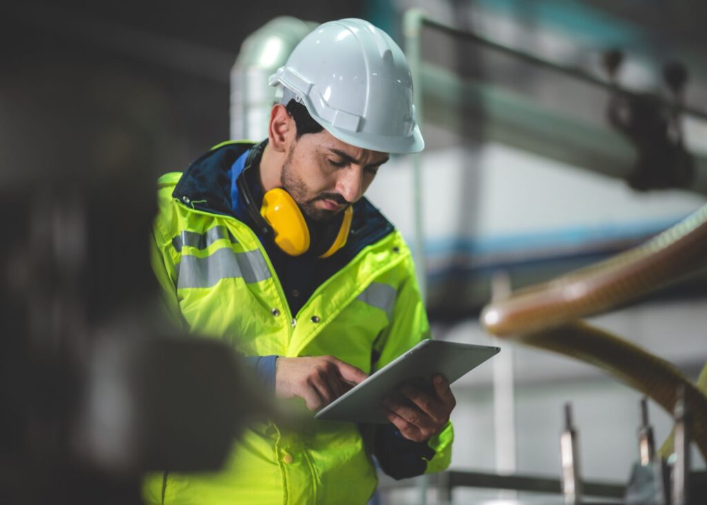 Man in hard hat, ear protection and yellow safety jacket uses an iPad to inspect equipment in a facitility